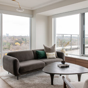 A bright, modern living room wrapped in large floor-to-ceiling windows that offer expansive views of a sprawling autumn forest and distant city skyline. The seating area features a sleek, curved dark grey-brown sofa styled with a vibrant emerald green pillow, sitting on a plush, patterned beige area rug. A dark wood, organic-shaped coffee table holds a stack of books, while a modern brass chandelier with flat white glass shades hangs from the coffered ceiling above light hardwood floors.