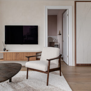 A modern living room featuring a mid-century inspired armchair with a dark wood frame and cream upholstery, placed on a plush textured rug next to a dark organic-shaped coffee table. In the background, a flat-screen TV is mounted above a custom floating media console with fluted wood details and rounded edges. To the right, a large built-in wardrobe showcases white stone-look doors with sleek brass pulls, framed in wood. An open doorway reveals a softly lit vanity area in the adjacent room.