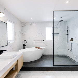 A spacious, modern luxury bathroom featuring large-format white marble-look wall tiles. The space includes a raised wet room area with dark grey floor tiles, housing a sleek white freestanding soaking tub with a wood caddy and a walk-in shower behind a single black-framed glass panel. Matte black fixtures are used throughout. To the left, a light wood double vanity features a thick white countertop, white vessel sinks, black faucets, and a large rectangular black-framed mirror.