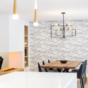 A bright, open-concept dining space viewed from a kitchen. The foreground features a blurred white quartz island illuminated by elegant gold and white cone pendant lights. In the background, a modern dining area showcases a thick wood dining table surrounded by dark grey chairs under a contemporary black and glass globe chandelier, set against a grey and white wavy patterned wallpaper. To the left, a sleek double-sided linear fireplace with an orange glow is built into a white room divider next to a light wood open shelving unit.