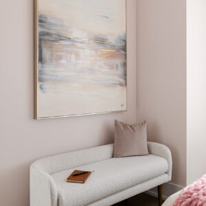 A cozy, modern bedroom corner featuring soft pastel pink walls and light wood flooring. A stylish white boucle upholstered bench with brass legs sits beneath a large, framed abstract painting with soft tones and gold leaf accents. The bench is elegantly styled with a dusty rose velvet pillow and a small brown leather journal. In the lower right foreground, a glimpse of a bed is visible, draped with a textured pink throw blanket.