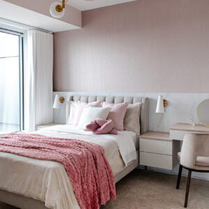 A bright, modern feminine bedroom featuring a soft pink upper wall above a sleek white marble-look lower accent wall. The bed has a light grey vertically tufted headboard and is elegantly styled with white and pink pillows alongside a textured pink throw blanket. Floating white nightstands with light wood tops flank the bed, seamlessly extending into a built-in vanity desk on the right. The space is illuminated by modern brass and white wall sconces and a unique geometric brass chandelier, with natural light pouring in from large windows draped in white curtains.