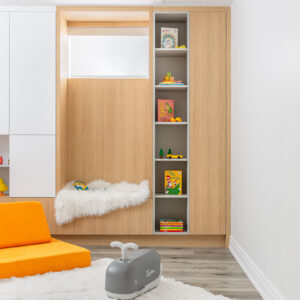A bright kids' playroom corner featuring custom built-in cabinetry. A cozy light wood-lined window seat niche is styled with a fluffy white throw, situated next to tall, narrow open grey shelves displaying children's books and small toys. A closed wood cabinet finishes the wall unit. The foreground features a vibrant orange modular play couch and a small grey ride-on toy resting on a soft white area rug over light grey wood-look flooring.