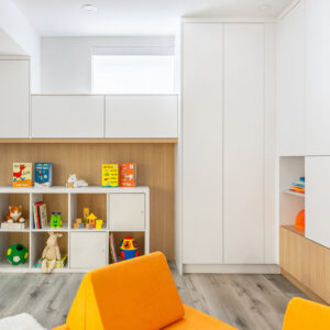 A brightly lit, modern children's playroom featuring sleek white floor-to-ceiling built-in cabinets and a wood-paneled wall alcove. A white cube storage unit filled with colorful toys, books, and stuffed animals sits neatly within the niche. In the foreground, vibrant orange modular play couch cushions rest on light grey wood-look flooring.