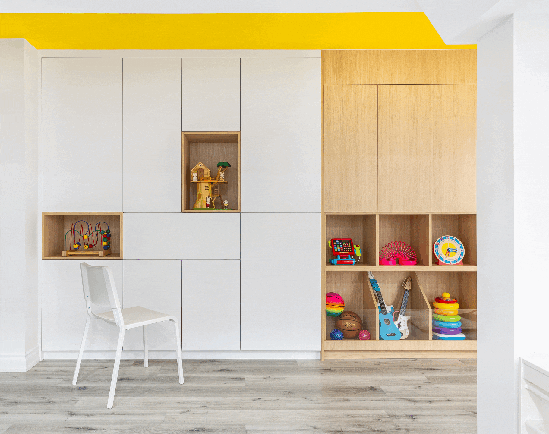 A modern children's playroom featuring a large wall of custom built-in cabinetry. The left side showcases sleek white cabinets with wood-lined display niches and an integrated drop-down desk with a modern white chair. The right side features light wood cabinets over open lower cubbies displaying colorful toys. A bright yellow painted ceiling adds a playful pop of color above the light grey wood-look flooring