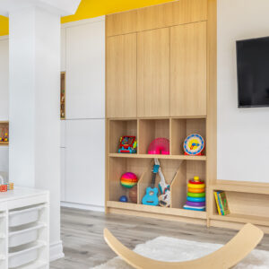 A bright and organized modern kids' playroom featuring a tall, custom light wood storage unit with closed upper cabinets and open lower cubbies displaying colorful toys like a toy guitar, balls, and stacking rings. A bold yellow painted ceiling accent adds a pop of color above. In the foreground, a curved wooden balance board rests on a soft white area rug over light grey wood-look flooring, with a white bin organizer visible to the left.