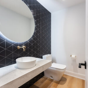 A striking modern powder room featuring a bold black geometric triangle tile accent wall. A thick, white floating stone vanity holds a round white vessel sink, elegantly paired with a wall-mounted brass faucet. A large, round backlit mirror adds a soft glow above the sink. The space is completed with a sleek modern white toilet, crisp white walls, and warm wood-look floor tiles.
