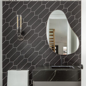 A modern powder room featuring a striking accent wall of black picket tiles laid diagonally with contrasting white grout, bordered by a strip of terrazzo at the top. An asymmetrical, organic-shaped mirror hangs above a dark grey vanity topped with a thick, black marble-look counter, integrated sink, and matte black faucet. A modern matte black double wall sconce with tubular filament bulbs illuminates the space next to a sleek, square-edged white toilet.