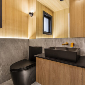 A sleek, contemporary powder room featuring a striking matte black one-piece toilet and a matching rectangular black vessel sink atop a floating wood vanity with a black countertop. A modern gold wall-mounted faucet adds a touch of luxury. The space is divided horizontally, with grey marble-look tile on the floor and lower half of the walls, contrasting with warm light wood paneling on the upper walls and ceiling. A large frameless mirror with warm integrated LED under-lighting reflects a black-framed window and modern black wall sconces.