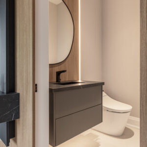 A peek into a modern, minimalist powder room featuring a sleek, dark grey floating vanity with a black stone countertop and a matte black faucet. A large round mirror is mounted against a warm, light wood accent panel that is beautifully illuminated by a vertical strip of LED backlighting. A modern white skirted toilet sits on light neutral floor tiles.