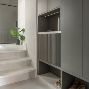A modern split-level entryway featuring custom dark grey built-in cabinetry. In the foreground, the multi-functional unit includes open shoe cubbies at the base, a textured fluted panel, and a backlit open drop-zone niche styling a minimal white vase. Short, light grey tiled stairs lead up to a landing that showcases matching floor-to-ceiling wardrobes with fluted detailing and a potted monstera plant.