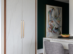 A modern interior featuring floor-to-ceiling custom cabinetry with white marble-look doors, warm wood framing, and sleek, elongated gold pull handles. To the right, a striking dark green accent wall displays a large framed abstract painting. In the foreground, a kitchen island with a light grey speckled stone waterfall countertop and textured grey bar stools is elegantly styled with a wooden bowl of green fruit resting on a round black tray.