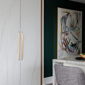 A modern interior featuring floor-to-ceiling custom cabinetry with white marble-look doors, warm wood framing, and sleek, elongated gold pull handles. To the right, a striking dark green accent wall displays a large framed abstract painting. In the foreground, a kitchen island with a light grey speckled stone waterfall countertop and textured grey bar stools is elegantly styled with a wooden bowl of green fruit resting on a round black tray.