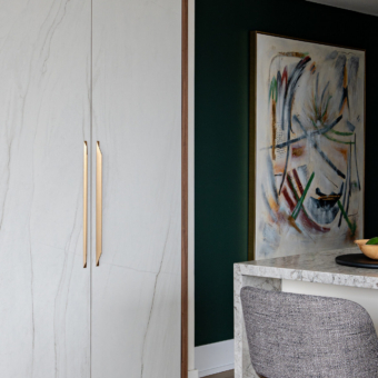 A modern interior featuring floor-to-ceiling custom cabinetry with white marble-look doors, warm wood framing, and sleek, elongated gold pull handles. To the right, a striking dark green accent wall displays a large framed abstract painting. In the foreground, a kitchen island with a light grey speckled stone waterfall countertop and textured grey bar stools is elegantly styled with a wooden bowl of green fruit resting on a round black tray.