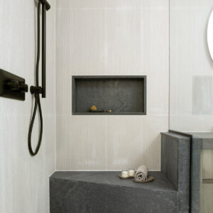 A close-up view of a modern walk-in shower featuring textured, off-white wall tiles and a light grey terrazzo floor. The design highlights a custom dark grey stone corner bench that seamlessly integrates with a half-wall dividing the shower from the adjacent vanity, complemented by a matching built-in stone shower niche. Sleek matte black fixtures, including a handheld shower wand on a slide bar, stand out against the light walls to complete the contemporary, spa-like aesthetic.