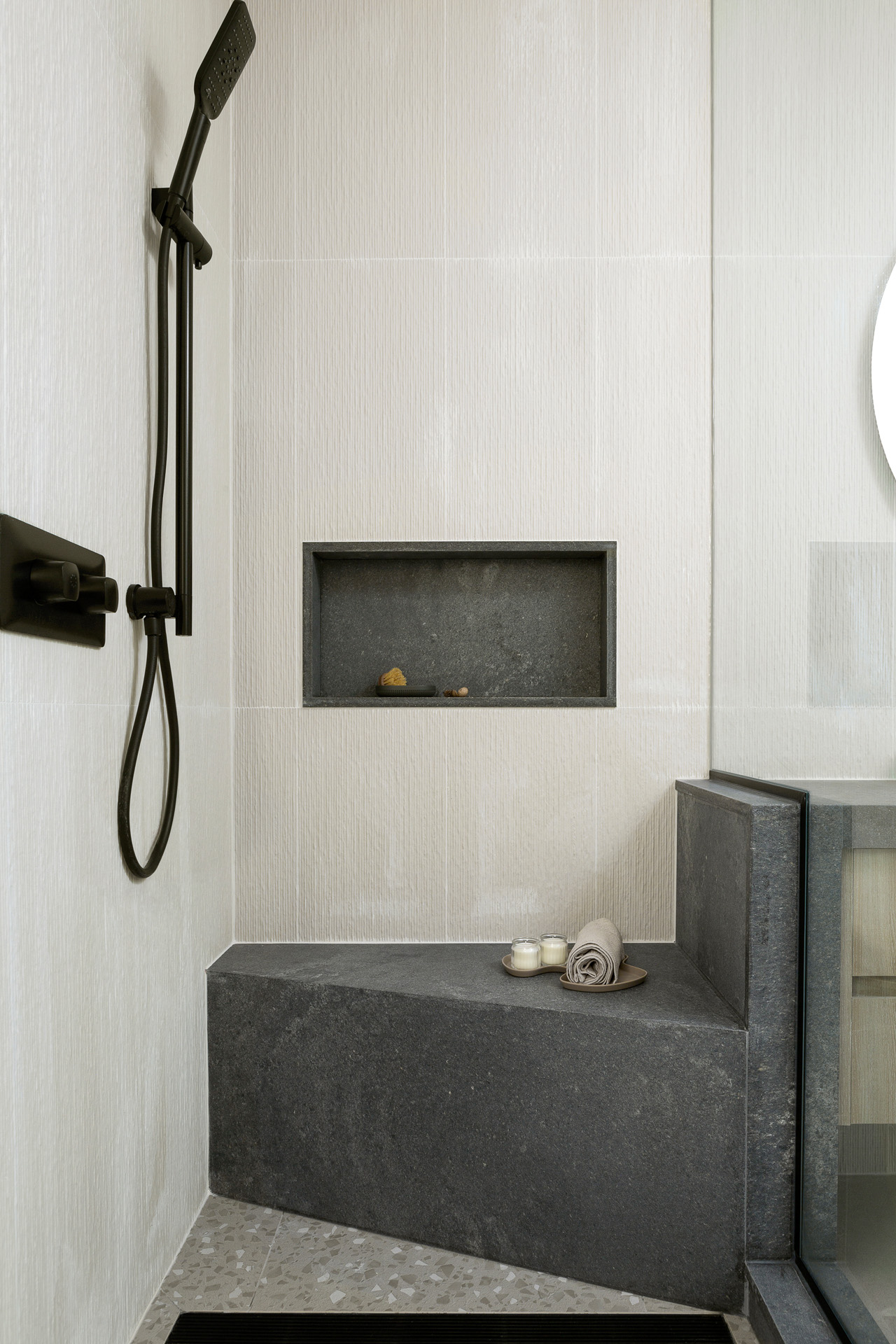 A close-up view of a modern walk-in shower featuring textured, off-white wall tiles and a light grey terrazzo floor. The design highlights a custom dark grey stone corner bench that seamlessly integrates with a half-wall dividing the shower from the adjacent vanity, complemented by a matching built-in stone shower niche. Sleek matte black fixtures, including a handheld shower wand on a slide bar, stand out against the light walls to complete the contemporary, spa-like aesthetic.