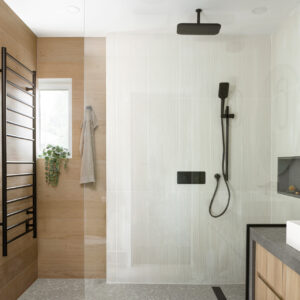 A modern walk-in shower featuring a frameless glass panel and a dark grey stone curb. The shower interior mixes warm, light wood-look wall tiles on the left with textured white tiles on the right, all grounded by a grey terrazzo tile floor. Sleek matte black fixtures, including a ceiling-mounted rain showerhead, a handheld wand, and a large heated towel rack, provide a striking contrast. A partial view of a matching light wood floating vanity with a dark grey countertop is visible to the right.