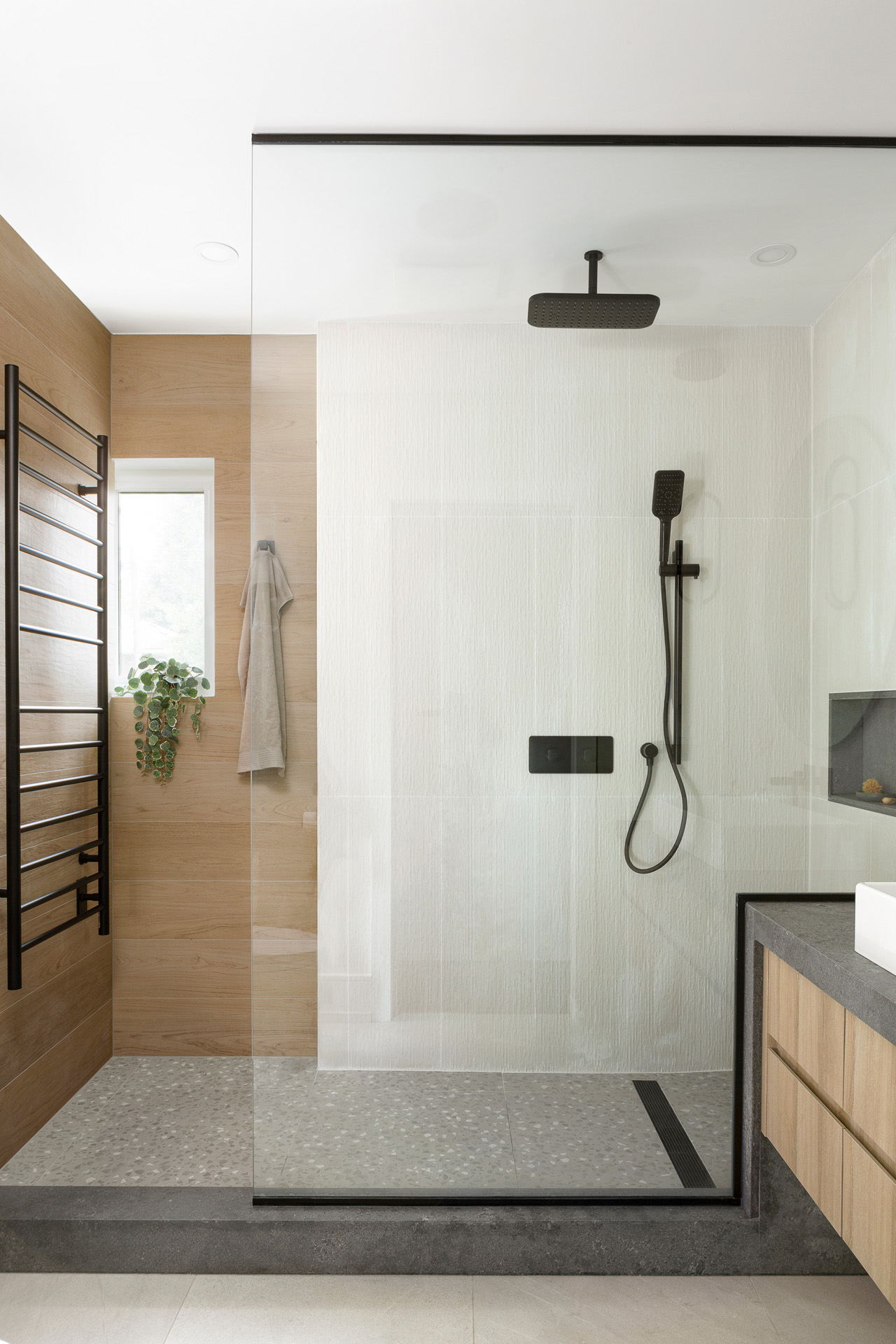 A modern walk-in shower featuring a frameless glass panel and a dark grey stone curb. The shower interior mixes warm, light wood-look wall tiles on the left with textured white tiles on the right, all grounded by a grey terrazzo tile floor. Sleek matte black fixtures, including a ceiling-mounted rain showerhead, a handheld wand, and a large heated towel rack, provide a striking contrast. A partial view of a matching light wood floating vanity with a dark grey countertop is visible to the right.