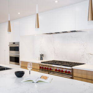 A bright, modern two-tone kitchen featuring flat-panel light wood lower cabinets and sleek white upper cabinets. A seamless white marble-look quartz forms the countertops and full backsplash. A professional stainless steel gas range with distinctive red knobs is centered on the back wall. The large white quartz island includes an under-mount sink, matte black faucet, and is illuminated by three elegant gold and white cone-shaped pendant lights.