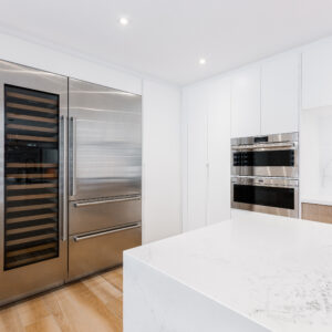 A sleek, modern kitchen featuring minimalist white flat-panel cabinetry and a thick white marble-look quartz waterfall island in the foreground. A prominent built-in stainless steel refrigerator sits side-by-side with a tall, glass-front wine column cooler. In the background, stainless steel wall ovens are seamlessly integrated into the white cabinets alongside a prep area with a matching quartz backsplash and light wood base cabinets.