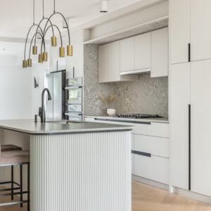 A bright, contemporary kitchen showcasing flat-panel white cabinetry accented with sleek, elongated black finger pulls. A unique terrazzo-style backsplash frames the stainless steel gas cooktop area. In the foreground, a prominent kitchen island features a distinctive rounded end with white fluted paneling and a light grey stone countertop equipped with a matte black faucet. Above the island hangs a striking modern linear chandelier with black arched arms and brass cylindrical shades. Light wood chevron flooring completes the sophisticated space.