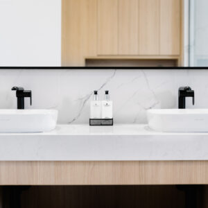 A close-up of a sleek, modern bathroom double vanity featuring a light wood base and a thick white marble-look countertop. Two white rectangular vessel sinks are paired with minimalist matte black faucets. A large rectangular black-framed mirror is mounted above a matching marble-look backsplash, reflecting a wooden slatted door in the background.