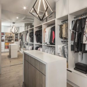 A spacious, modern walk-in closet featuring extensive custom white built-in open shelving and hanging racks filled with neatly organized clothing. In the foreground, a sleek central island features a thick white countertop and a warm wood-grain base. A striking black geometric diamond-shaped LED pendant light hangs from the ceiling. The back wall consists of floor-to-ceiling mirrors that reflect the closet's depth, the light wood flooring, and additional illuminated display shelving for accessories.