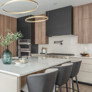 An angled view of a modern kitchen featuring a large island with a white quartz countertop and three curved black leather counter stools. Two contemporary gold halo pendant lights hang elegantly above. The back wall showcases a striking matte black range hood, a seamless white quartz slab backsplash with a black pot filler, and a stainless steel gas range, all framed by sleek, flat-panel light wood cabinetry.