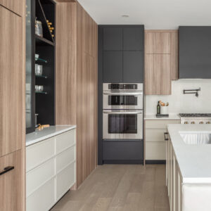 A modern kitchen featuring a stainless steel double wall oven integrated into a sleek matte black floor-to-ceiling cabinet. To the left, flat-panel light wood cabinetry frames a custom built-in bar area with glass shelving, a black interior, and light beige lower drawers. Light oak hardwood flooring and the edge of a white quartz kitchen island complete the contemporary space.