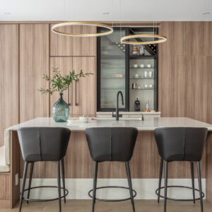 A bright, modern kitchen featuring a large light-wood paneled island with a sleek white countertop and three black leather counter stools. Two contemporary gold halo pendant lights hang overhead. The background showcases seamless floor-to-ceiling wood cabinetry that integrates a custom black glass-front bar and coffee station. To the left, a built-in banquette with fluted wood detailing connects seamlessly to the island, creating a cozy dining nook alongside a white tulip table.