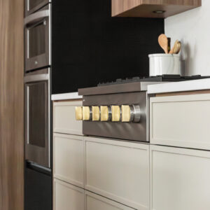 A close-up view of a modern kitchen featuring a stainless steel gas range with distinctive square brass knobs. The cooktop is set above sleek, light beige flat-panel lower drawers, sharply contrasted by a matte black background and a built-in stainless steel double wall oven to the left.