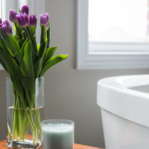 A close-up view of a bathroom styling detail featuring a rectangular clear glass vase filled with fresh, vibrant purple tulips on a small wooden accent table. Next to the flowers sits a light green scented jar candle in a clear glass container. In the blurred background, a white freestanding soaking bathtub sits near a large window, creating a serene and spa-like atmosphere.