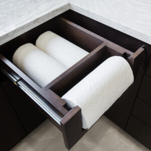 An overhead view of an open custom wooden drawer in a modern kitchen, showcasing an integrated paper towel dispenser and storage system. The dark espresso wood drawer features a built-in rod holding an active roll of paper towels at the front, with a rear compartment organized to store two additional backup rolls. The unit is installed into dark base cabinetry beneath a light grey veined stone countertop.
