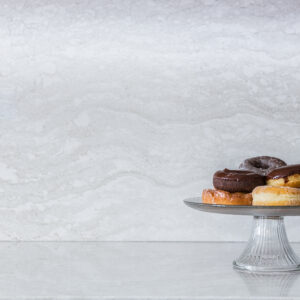 A clear glass pedestal cake stand filled with an assortment of donuts—including chocolate frosted, glazed, and powdered varieties—sitting on a polished white marble countertop. The background consists of a matching seamless full-height marble slab backsplash featuring elegant, soft grey wavy veining patterns, leaving ample negative space on the left.
