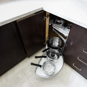 An overhead view of a modern kitchen's blind corner base cabinet with its door open, showcasing an innovative kidney-shaped pull-out organizer system. The multi-tiered white shelving unit smoothly glides out from the deep corner space to provide easy access to a collection of stored stainless steel pots and pans. The surrounding cabinetry features a dark espresso wood finish with modern chrome pulls, set beneath a seamless white quartz countertop with an under-mount sink visible on the left.