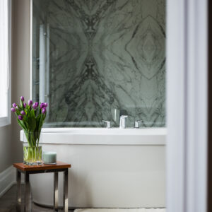 A luxury primary bathroom featuring a white freestanding soaking bathtub set against a dramatic, full-height bookmatched marble accent wall with symmetrical grey veining. A modern chrome deck-mounted faucet sits on the edge of the tub. In the foreground, a small wooden side table with a polished chrome base holds a glass vase of vibrant purple tulips and a scented candle near a plush white bath mat on glossy grey tile flooring.