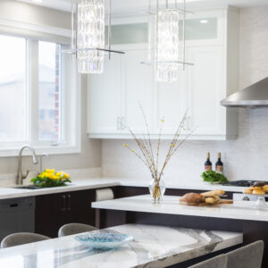 A bright, luxury kitchen showcasing two modern chrome and crystal cylinder pendant lights hanging over a multi-level island. The lower tier features a polished white and grey marbled waterfall dining table lined with grey upholstered chairs. The upper island tier has a dark wood base with a white quartz countertop styled with fresh bread and a vase of yellow flower buds. In the background, dark wood base cabinets meet white shaker upper units along a light stone backsplash, complete with a stainless steel sink under a large window.