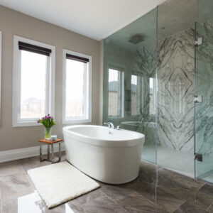 A spacious luxury primary bathroom featuring a freestanding white soaking tub and a massive seamless glass walk-in shower. The shower interior boasts striking, full-height bookmatched marble accent walls with symmetrical grey veining. Glossy grey marble-look floor tiles run throughout the space, complemented by greige walls and three large windows with dark blinds that fill the room with natural light. A plush white bath mat lies beside the tub, along with a small wooden side table styled with fresh purple tulips.
