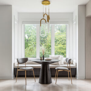 A bright, modern breakfast nook centered around a large bay window with lush green views. The inviting space features a custom built-in window bench with a dark base and light beige upholstered seating, paired with a modern round black dining table with a textured conical base. Two sleek grey chairs with gold legs sit opposite the bench, while a unique interlocking brass and white pendant light hangs above, completing the elegant design.