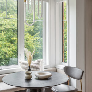 A close-up view of a stylish modern breakfast nook featuring a round black dining table with a textured wood-grain top and a ribbed conical base. Next to it sits a sleek grey upholstered chair with slender gold legs. A built-in window bench with a light beige cushion and dark cabinetry base wraps around the bright corner windows, offering views of lush greenery. Minimalist ceramic decor and dried pampas grass style the table, while a unique brass and white loop pendant light hangs above.