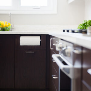 A close-up view of a modern kitchen corner featuring dark espresso flat-panel wood cabinets with slim silver handles. A clever built-in paper towel holder niche is integrated seamlessly into the upper portion of a base cabinet drawer. The clean white quartz countertops are styled with a vase of bright yellow flowers near a stainless steel gooseneck faucet on the left, and a partial view of a gas cooktop, stainless steel wall oven, fresh greens, and wine bottles on the right.