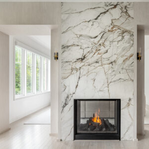 A luxurious modern living space featuring a stunning double-sided fireplace that acts as a central room divider. The floor-to-ceiling fireplace surround is clad in striking white marble with dramatic dark grey and gold veining. A warm fire burns within the black glass firebox, providing a see-through view to the adjacent room. The structure is flanked by two open passageways leading into a bright, window-filled space. The textured light neutral walls are accented with elegant brass and black glass sconces above light hardwood floors.