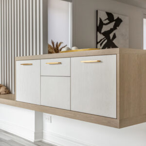 A close-up view of a custom modern floating console cabinet. The design features a light wood-grain frame wrapping around white flat-panel doors and drawers, beautifully accented with sleek gold hardware. The built-in unit extends from a wooden bench area, set against a crisp white wall with vertical slatted detailing and a glimpse of abstract black and white art in the background.