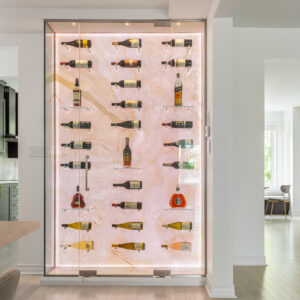A close-up view of a custom floor-to-ceiling glass-enclosed wine display in a modern home. The unit features a stunning backlit translucent stone back wall, resembling warm onyx or marble, which beautifully illuminates the wine bottles resting on minimal metal pegs and small glass shelves. This luxury built-in feature is seamlessly integrated into crisp white walls above light oak hardwood flooring.