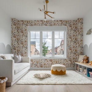 A bright, modern kids' playroom featuring a central window framed by whimsical, earth-toned botanical and animal patterned wallpaper. To the left, a comfortable white sofa sits against a wall with painted grey scalloped details. On the right, a low white cubby bench provides toy storage and seating. The cozy space is tied together with a fluffy white area rug, a textured mustard and white pouf, light wood flooring, and a modern brass sputnik globe chandelier.