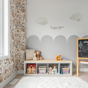 A cozy corner of a modern kids' playroom featuring a low white cubby storage bench with a light wood top, organized with wire toy baskets, a small dollhouse, and stuffed animals. The wall behind the bench is painted with a playful grey scalloped design, adorned with cloud-shaped mirrors and a wire 'happy place' sign. To the left, an accent wall showcases earth-toned botanical wallpaper, all resting on a fluffy white rug and light wood flooring next to a wooden chalkboard easel.