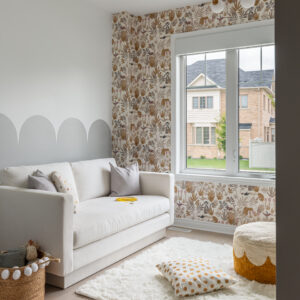 A cozy corner of a bright, modern kids' playroom featuring a clean-lined white sofa set against a wall with a painted light grey scalloped design. The adjacent wall surrounding a large window showcases whimsical, earth-toned botanical and animal wallpaper. The space is beautifully styled with a fluffy white area rug, a polka-dot floor pillow, a textured mustard and white pouf, and a woven toy basket with white pom-poms on light wood flooring.