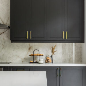 A close-up view of a sophisticated modern kitchen featuring matte black shaker-style cabinetry accented with sleek gold bar pulls. The design showcases a crisp white countertop seamlessly transitioning into a full-height light stone backsplash with warm, natural veining. The counter is elegantly styled with a black two-tier snack tray, a glass cookie jar, and minimalist decor, framed by a partial view of a white kitchen island in the foreground.