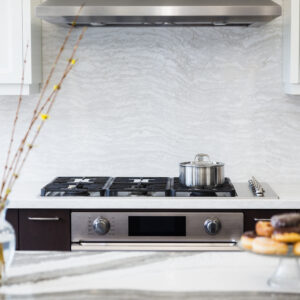 A close-up view of a modern kitchen cooking station featuring a stainless steel gas cooktop with a metal pot resting on a burner, paired with a built-in stainless steel wall oven below. A seamless, full-height light grey quartzite or marble slab backsplash with a subtle wavy vein pattern runs up to a matching stainless steel range hood. The workspace is framed by dark wood base cabinets and white upper cabinetry, with a blurred foreground showing a veined kitchen island styled with a glass vase of yellow flower buds and a glass stand of donuts.