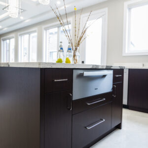 A low-angle view of a modern kitchen island with a dark espresso wood base and a thick white marbled quartz countertop. A built-in stainless steel Miele speed oven or microwave drawer is integrated seamlessly into the center of the cabinetry, surrounded by flat-panel drawers and cupboards with sleek silver handles. The countertop is styled with a vase of yellow flower buds and glass water bottles. Two crystal cylinder pendant lights hang from a recessed tray ceiling above, with large windows and bright tile flooring completing the background.